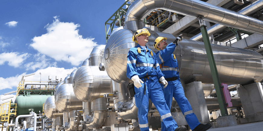 Two oil and gas workers in blue coveralls and yellow hard hats walking on site, talking.