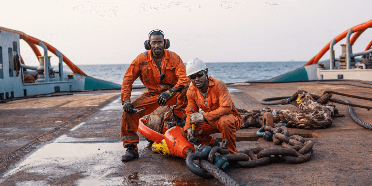 Two offshore workers on the deck of an offshore vessel squatting over a large chain smiling for the camera.